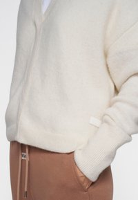 Crème gebreide cardigan met V-hals, geribbelde manchetten en zoom. Zachte textuur. Klein merkje op de onderkant rechts. Gemaakt over een paar bruine joggingbroek.
