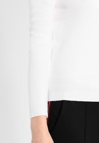 Bílý žebrovaný pletený svetr s dlouhým rukávem a přiléhavým střihem. Manžeta má kontrastní strukturovaný okraj. Kombinováno s černými kalhotami.