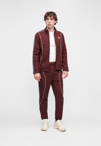 Bordó színű melegítő fehér csíkozással, cipzáras felsővel és húzózsinóros nadrággal. A modell szűk fehér alsóinget és világos sportcipőt visel.