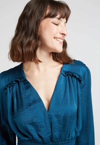 Robe en tissu plissé teal avec un décolleté en V profond, des accents de manches à volants et une taille élastique pour une silhouette ajustée.