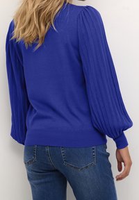Niebieski sweter z prążkowanymi bufkami na rękawach, o dzianinowej fakturze i okrągłym dekoltem. Połączony z dżinsami.