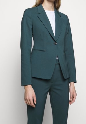 Mujer con un blazer entallado verde oscuro con un solo botón sobre una camisa blanca y pantalones verde oscuro a juego.