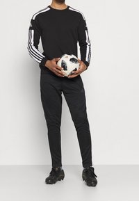 Maglietta sportiva nera a maniche lunghe con strisce bianche sulle maniche, pantaloni neri e scarpette nere, mentre tiene in mano un pallone da calcio a fantasia.