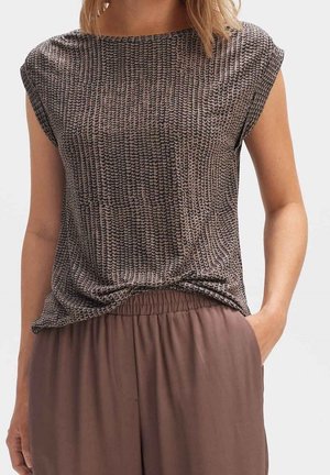 Femme portant un haut marron et noir à manches courtes et motifs, légèrement rentré dans un pantalon marron ample taille haute, une main dans la poche.