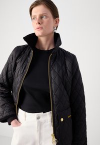 Zwarte gewatteerde jas met een hoge kraag, gouden ritssluiting en zakaccenten. Gedragen over een zwart geribbeld topje en witte broek.