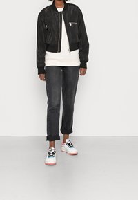 Chaqueta bomber negra corta con bolsillos con cremallera, llevada sobre una sudadera blanca, combinada con vaqueros oscuros remangados y zapatillas de colores.