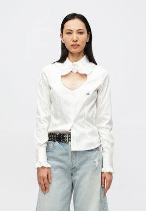 Camisa branca de botão com gola franzida, recorte em forma de coração na frente e detalhe bordado. Combinada com jeans azul claro desfiados e um cinto preto.