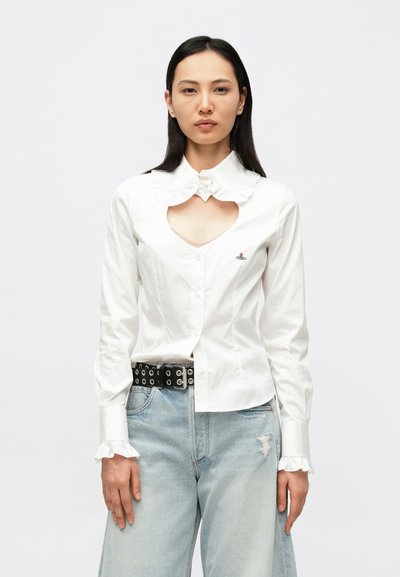 Weiße Knopfleiste Bluse mit gerafftem Kragen, herzförmigem Ausschnitt vorne und besticktem Detail. Kombiniert mit hellblauen, verwaschenen Jeans und einem schwarzen Gürtel.
