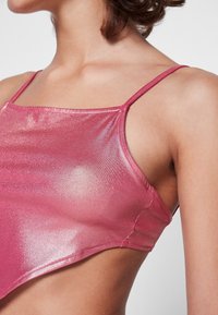 Top corto metálico rosa con tirantes finos, que presenta una textura suave y ligeramente brillante y un diseño ajustado que resalta los hombros.