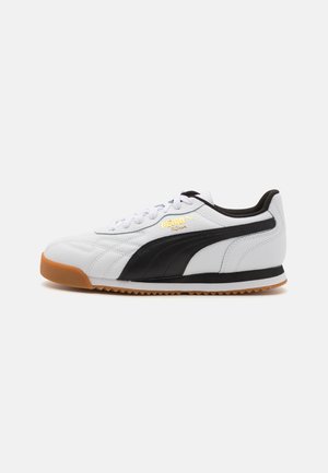 White leather sneaker with black stripes and textured accents. Features a gum rubber sole and embossed branding in gold. Rounded toe shape.