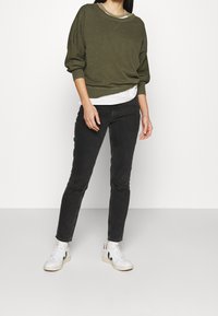 Olijfgroene sweatshirt met uitgezakte schouders, gelaagd over een wit shirt. Zwarte skinny jeans en witte sneakers met zwarte accenten.