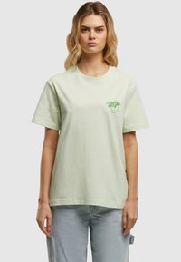 Lichtgroene katoenen t-shirt met korte mouwen, voorzien van een kleine groene plantafbeelding op de linkerkant van de borst. Casual pasvorm, zachte textuur.