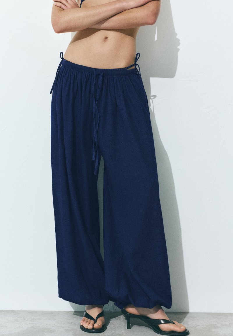 Personne portant un pantalon ample bleu marine avec des cordons et des sandales noires à talons, debout contre un mur blanc, les bras croisés.