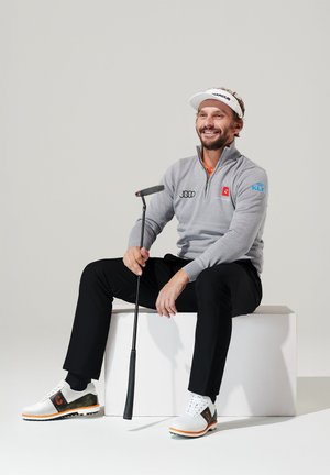 Gray zippered long-sleeve sweater, black pants, white golf shoes with orange accents. Man sitting on a white block, holding a putter.
