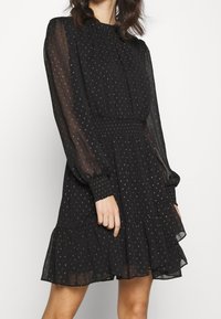 Vestido negro de tela transparente, con cintura fruncida, mangas largas y dobladillo con volantes. Pequeños patrones geométricos rosas en toda la prenda.