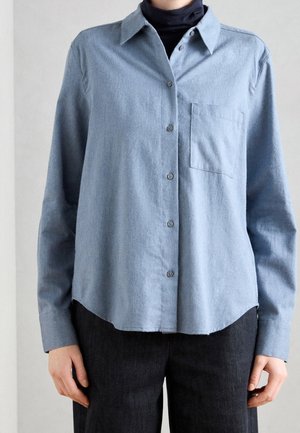 Person wearing a light blue button-up shirt over a black turtleneck, paired with dark pants, standing against a plain background.