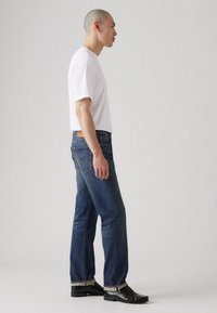 Camiseta blanca ajustada de hombre, jeans azules con rodillas desgastadas, costuras en contraste y parche de cuero marrón. Usada con mocasines negros.