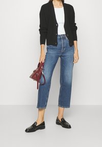 Femme portant un jeans bleu cropped, un cardigan noir à boutons, un haut blanc, des mocassins noirs à clous, tenant un petit sac à main bordeaux sur fond blanc.