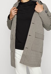 Abrigo de pata de gallo en beige y negro, con grandes bolsillos frontales, botones y un corte relajado. Suéter negro debajo. Pantalones blancos.