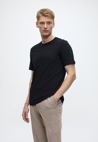 Schwarzes kurzärmeliges T-Shirt mit rundem Ausschnitt und glatter Textur, getragen zu hellbraunen gemusterten Hosen, Hände in den Taschen.