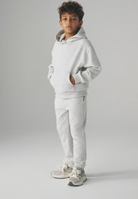 Chłopiec stoi z rękami w kieszeniach, ubrany w jasnoszary hoodie, pasujące dresy oraz beżowe sneakersy na jednolitym tle.