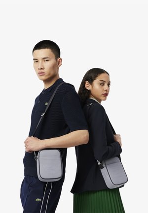 Borsa a tracolla rettangolare grigia con logo impresso, tracolla regolabile e dettagli perforati. I modelli indossano outfit casual in colori contrastanti.