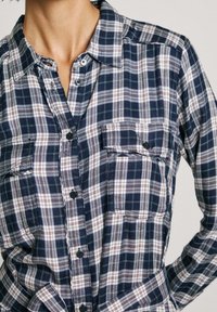 Camicia flanella a scacchi blu navy e bianca con design a bottone, due tasche frontali e un tessuto morbido e texture.