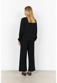 Woman standing facing a white wall wearing a black long-sleeve top, wide-leg black pants, and studded dark brown flat shoes.