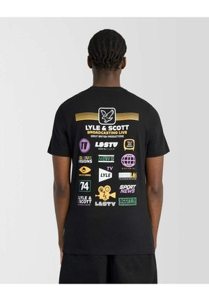 Mand iført sort t-shirt med farverige tv-kanal-logoer og tekst relateret til Lyle & Scott Broadcasting på ryggen.