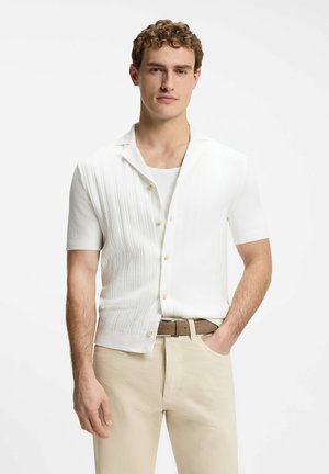 Homme aux cheveux bouclés portant une chemise blanche à manches courtes sur un débardeur blanc et un pantalon beige avec une ceinture marron, main dans la poche.