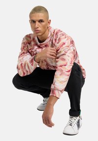 Langarm-Sweatshirt in Pink und Creme mit einem wirbelnden Muster, kombiniert mit schwarzen Hosen und weißen Sneakers mit schwarzen Akzenten.