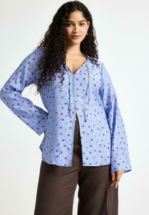 Donna con capelli lunghi e ricci che indossa una camicetta azzurro chiaro con piccoli motivi floreali e pantaloni marrone scuro, in piedi con una mano sul fianco.