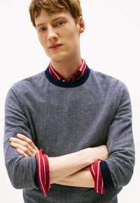Giovane uomo con le braccia incrociate che indossa un maglione a motivi blu navy sopra una camicia a righe rosse, contro uno sfondo semplice e chiaro.