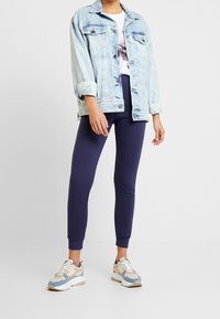 Jeansjacka i ljusblått, ribbade muddar, bärs över en vit T-shirt. Marinblå leggings och mångfärgade träningsskor fulländar outfiten.