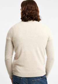 Vaaleanbeige neulepaita, joka on valmistettu pehmeästä, tekstuurista kankaasta. Neulotut yksityiskohdat pitkissä hihoissa ja resorikäsineissä, takanäkymä. Denim-alusvaatteet näkyvät.