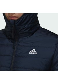 Detailní záběr na osobu oblečenou v tmavě modré péřové bundě značky Adidas, zapnuté až ke krku, s viditelným logem značky na hrudi.
