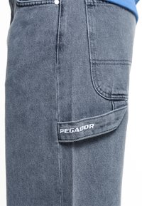 Närbild av blå denimbyxor med en sidoficka som har en klaff broderad med ordet "PEGADOR" i vit tråd.