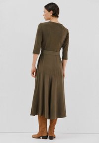 Oliwkowa sukienka do kolan wykonana z miękkiego materiału, z paskiem w talii i rękawami o długości trzech kwartałów, w połączeniu z brązowymi wysokimi butami do kolan.