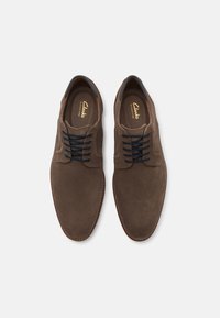 Brune semskede sko med en glatt tekstur, med en spiss tupp, mørke snørebånd og en sydd brun såle. Clarks-logoet er synlig inni.