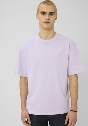 Jeune homme aux cheveux blonds courts portant un t-shirt ample uni violet clair et un jean foncé, debout devant un fond neutre.