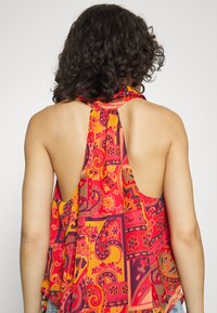 Top leve e sem mangas com decote tipo halter em vermelho e laranja, apresentando padrões intrincados de paisley e flores. Design com costas abertas e detalhe de laço no pescoço.