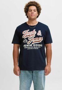 Tamnoplava pamučna majica s bijelim i crvenim natpisima "Jack & Jones" i "Denim Goods". Uparen sa svijetloplavim trapericama.