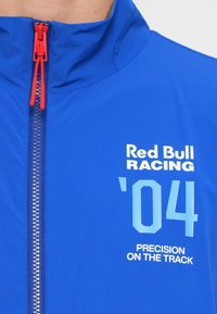 Blauwe racejas gemaakt van lichtgewicht materiaal. Voorzien van een hoge kraag, een volledige ritssluiting met een rode trekker en bedrukt met witte en lichtblauwe graphics.