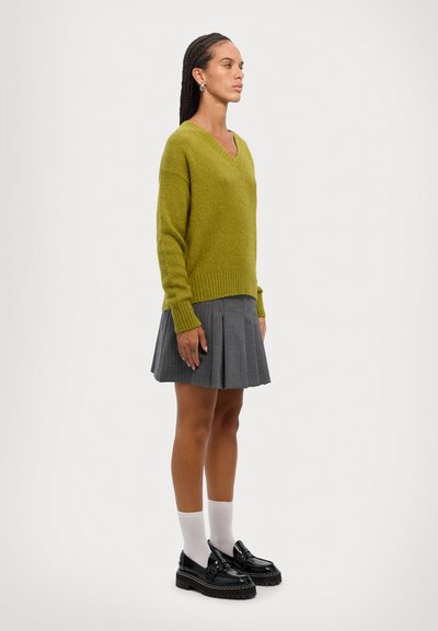 Un pull en V vert tricoté, une jupe grise plissée, des chaussettes blanches et des mocassins noirs. Le pull a un ourlet côtelé ; la jupe présente un léger motif à carreaux.