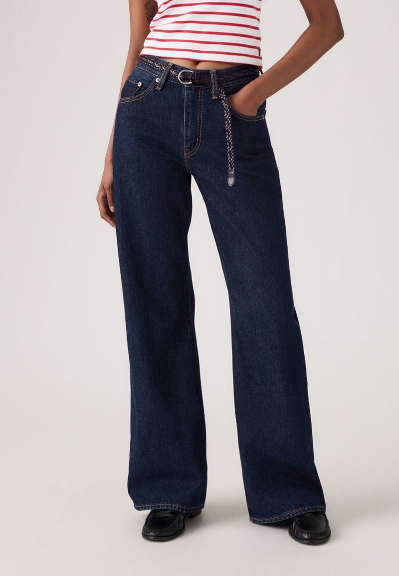 Femme portant un jean bleu foncé taille haute à jambes larges avec une ceinture tressée, un crop top rayé et des chaussures noires, debout devant un fond uni.