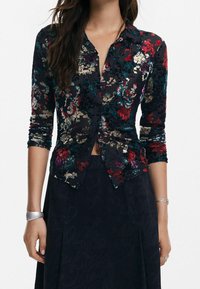 Long-sleeve shirt in floral velvet fabric, featuring a dark navy background with red, green, and cream patterns. Buttoned front and fitted design.