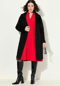 Cappotto nero con cappuccio, chiusura a un solo bottone, indossato sopra un vestito rosso. Stivali neri fino al ginocchio e occhiali da sole neri tenuti in una mano.