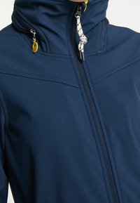 Námořnická modrá softshell bunda s vysokým límcem, předním zipem a žlutými akcenty. Má texturovanou tkaninu s jemným leskem.