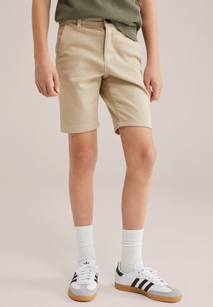 Pois, kes kannab beežikaid lühikesi pükse, valgeid Adidase tossusid mustade triipudega ja valgeid sokke, seisab ühevärvilise tausta ees.