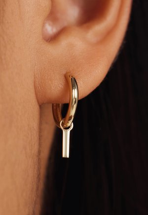 Gouden hoepeloorbel met een stijlvolle, gepolijste afwerking en een verticale staafaccent dat aan de onderkant is bevestigd. Close-up zicht tegen de huidskleur.